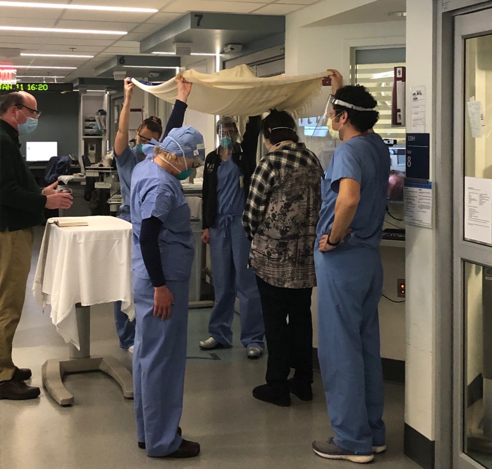 In the hallway of the ICU, four hospital staff in scrubs hold a chuppah over the head of a woman, while the rabbi officiates using a bedside tray as a table.